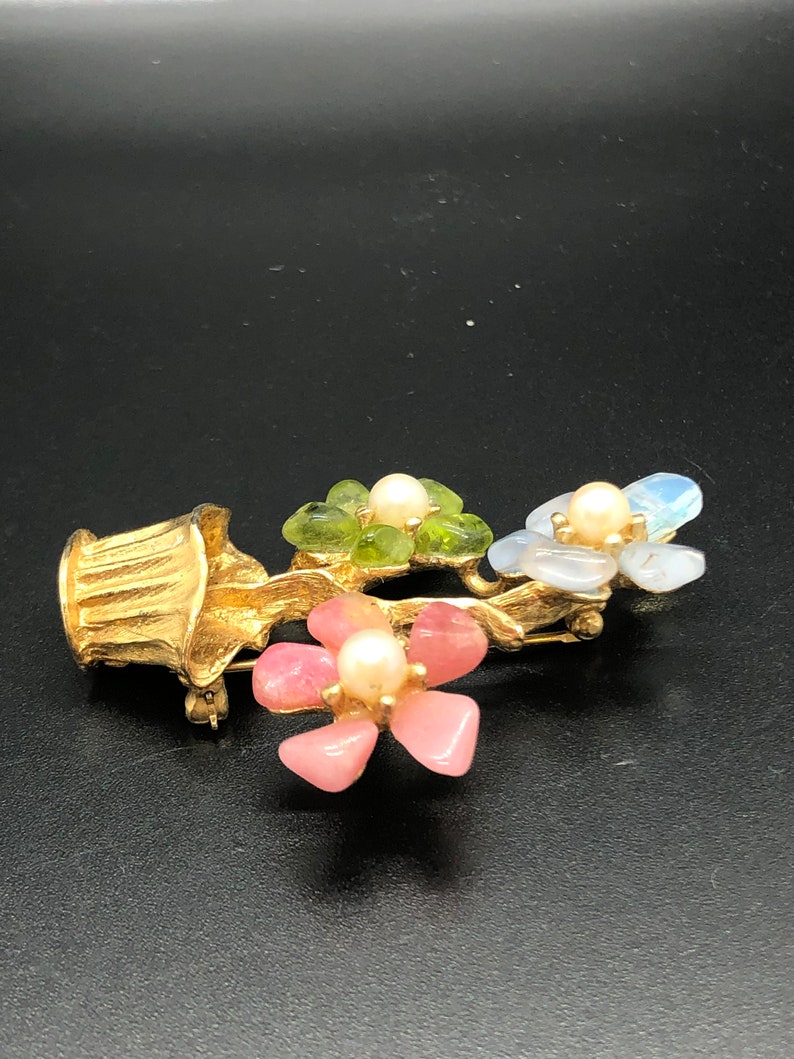 Puede incluir: Broche floral dorado con tres grupos de flores, cada uno con un centro de perla y p&eacute;talos de piedra de colores. El broche tiene un acabado dorado texturizado y un cierre de alfiler. Las flores son rosas, verdes y azules.