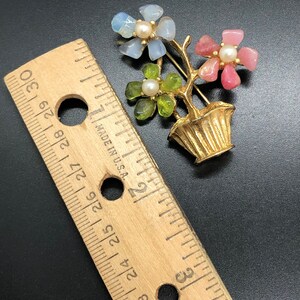 Puede incluir: Broche de metal dorado con forma de arreglo floral en maceta. Las flores son de piedras azules, rosas y verdes, cada una con un centro de perla. La broche mide aproximadamente 5 cm de alto.