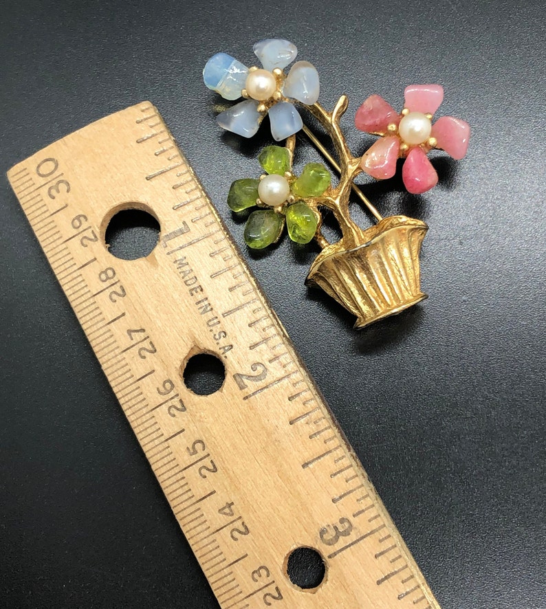 Puede incluir: Broche de metal dorado con forma de arreglo floral en maceta. Las flores son de piedras azules, rosas y verdes, cada una con un centro de perla. La broche mide aproximadamente 5 cm de alto.
