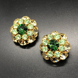 May include: A pair of vintage clip-on earrings. Each earring features a central, large, round, dark green rhinestone surrounded by smaller light green and clear rhinestones, set in a gold-tone metal flower shape.