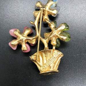 Puede incluir: Broche vintage dorado con dise&ntilde;o floral. El broche presenta tres grupos de flores en rosa, azul y verde. Las flores est&aacute;n colocadas en una maceta dorada con el texto "SWOBOON, INC."