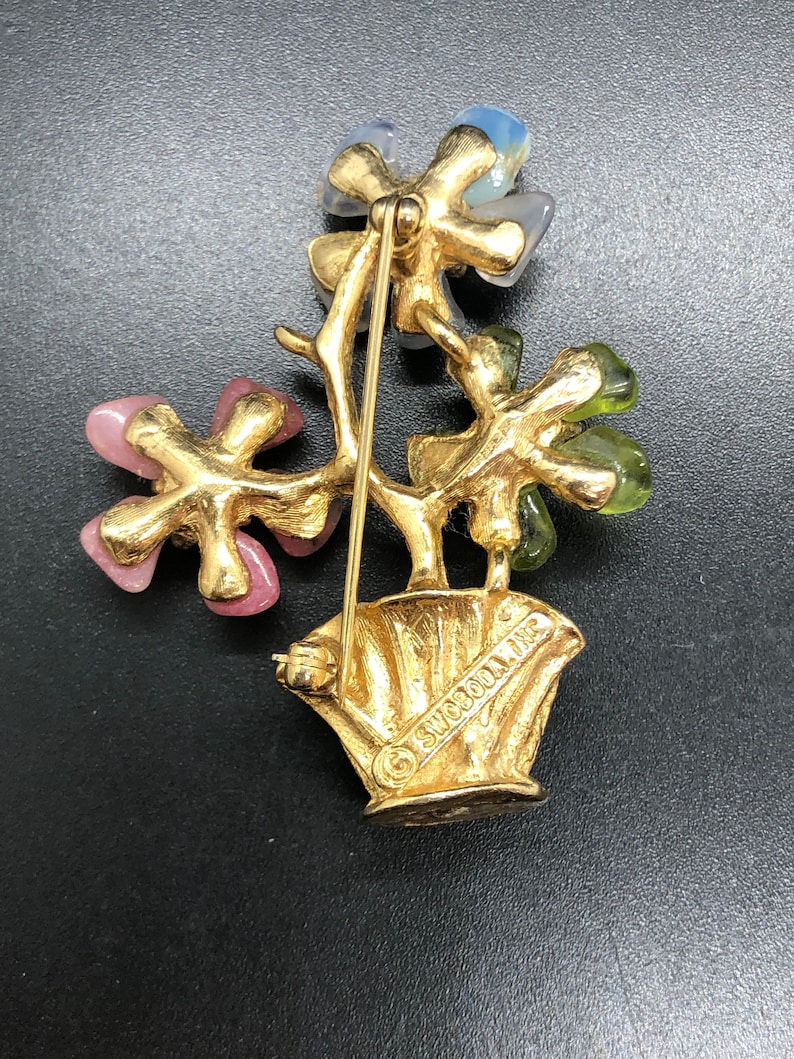 Puede incluir: Broche vintage dorado con dise&ntilde;o floral. El broche presenta tres grupos de flores en rosa, azul y verde. Las flores est&aacute;n colocadas en una maceta dorada con el texto "SWOBOON, INC."