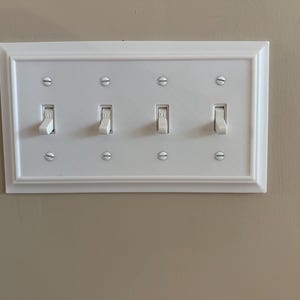 May include: A white, rectangular light switch plate with four toggle switches. The plate has a decorative border and is mounted on a beige wall. Each switch is in the on or off position.