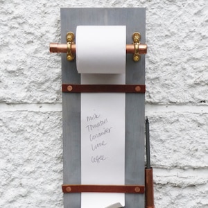 May include: A grey wooden memo board with a copper pipe and gold hardware holding a white paper roll. The paper has a handwritten grocery list that reads: "Milk, Tomatoes, Coriander, Lime, Coffee". A copper pencil holder with a grey pencil is on the right side of the board.