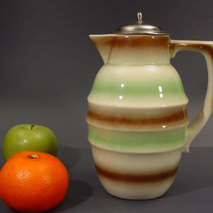 May include: A striped ceramic pitcher with a silver lid. The pitcher has brown and green stripes and a cream-colored background.