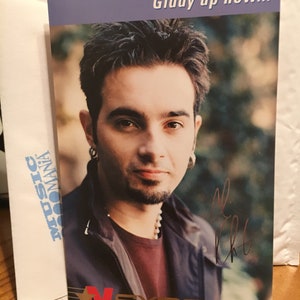 May include: A close-up of a man with dark hair and a goatee, wearing a black jacket over a red shirt. He is looking at the camera and has a serious expression. The image is from a magazine or promotional card and includes the text "Giddy up now..." and "NSYNC".