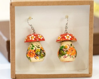 May include: A pair of wooden mushroom earrings with red and white tops and floral designs in orange, yellow, and green. The earrings are displayed in a brown cardboard box.
