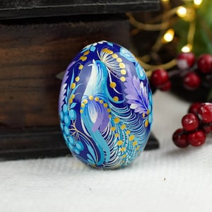 May include: A blue and gold hand-painted wooden egg with a peacock design. The egg is decorated with intricate details and vibrant colors.