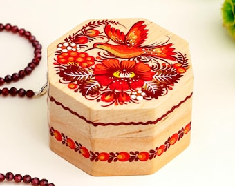 May include: A wooden, octagonal box with a hand-painted floral and bird design in red, orange, and green. A beaded necklace with small, dark red beads is draped nearby. Green flowers are in the background.