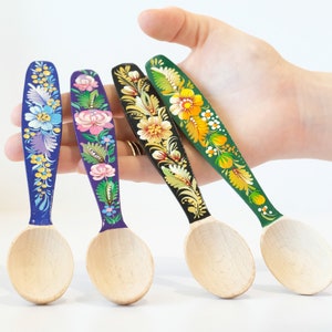 May include: Four wooden spoons with hand-painted floral designs. The spoons are painted in shades of blue, purple, black, and green. The designs feature flowers, leaves, and swirls.