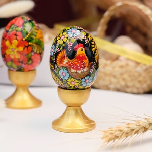 Op de afbeelding: Twee handgeschilderde houten paaseieren op gouden standaards. Eén ei heeft een rode haan met zwarte en gele accenten, omringd door kleurrijke bloemen. Het andere ei heeft een bloemmotief met rode, gele en blauwe bloemen.