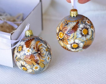 May include: Two hand-painted Christmas ornaments with a floral design. The ornaments are a light gray color with white and yellow flowers. The ornaments are in a white box with white ribbon.