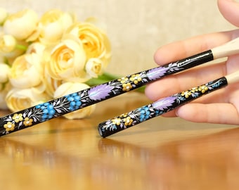 May include: Two black cigarette holders with a hand-painted floral pattern featuring yellow flowers, blue berries, and purple leaves. Each holder has a light-colored mouthpiece and a slender, cylindrical body with a glossy finish.