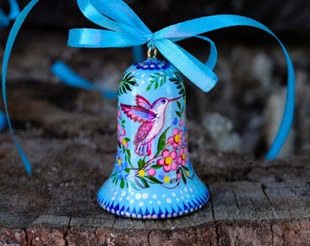 Puede incluir: Un adorno de campana azul con un colibrí pintado y un diseño floral. La campana está atada con una cinta azul.