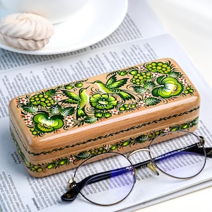 Puede incluir: Un estuche de madera para gafas con un diseño floral pintado a mano en verde y blanco, con colibríes. El estuche está sobre un periódico junto a una taza de café y unas gafas redondas.