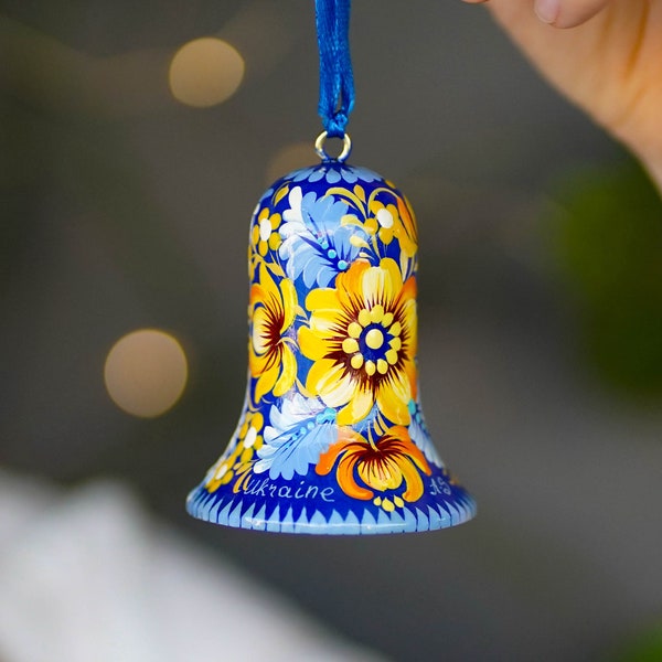 以下が含まれることがあります： 黄色と白の花柄の青いベルのオーナメント。ベルには花が描かれており、「ウクライナ」という言葉が書かれています。