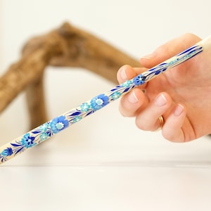 Puede incluir: Una varita larga, delgada y cilíndrica con una base de color crema, decorada con un patrón repetitivo de flores de color azul vibrante y azul claro, hojas azul oscuro y tallos finos que envuelven toda la longitud del objeto de madera cónico.