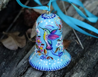 May include: A blue bell ornament with a painted hummingbird and floral design. The bell has a white ribbon attached to it.