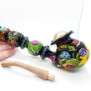 May include: A black, hand-painted smoking pipe featuring colorful ladybugs, green leaves, and floral patterns. The pipe has a long, segmented stem and a metallic bowl cover. A separate, curved, light-colored wooden stem piece is included.