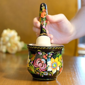 Puede incluir: Un mortero y un pilón de madera negro con diseños florales coloridos. El pilón está siendo sostenido por una mano y está a punto de ser usado para moler algo.
