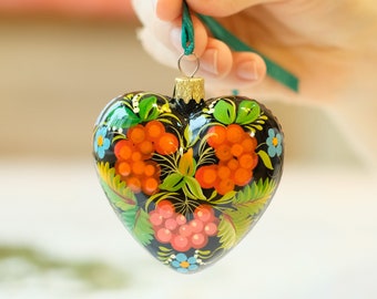 May include: A heart-shaped Christmas ornament painted with a black background and colorful floral designs. The ornament features red berries, green leaves, and blue flowers.