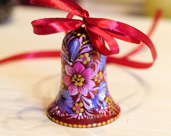 May include: A hand-painted wooden bell with a red ribbon. The bell is decorated with pink, purple, and gold flowers. The bell has the word "Ukraine" written on it.