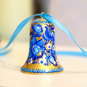 Puede incluir: Una campana dorada con dise&ntilde;os florales azules y blancos pintados en ella. La campana est&aacute; atada con una cinta azul claro.