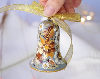 May include: A hand-painted ceramic bell ornament with a floral design in shades of brown, yellow, and white on a light blue background. The bell is tied with a gold and white ribbon.