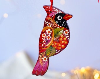 May include: A hand-painted wooden cardinal Christmas ornament with red, pink, green, and gold details. The bird is decorated with white flowers and red berries.