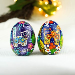May include: Two blue Easter eggs with hand-painted designs of cats and flowers. The eggs are decorated with bright colors, including yellow, orange, red, green, blue, and purple.