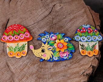 May include: Three wooden ornaments painted with colourful flowers and designs. The ornaments are shaped like a mushroom, a hedgehog, and a mushroom. The ornaments are on a wooden surface.