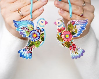 Könnte beinhalten: Zwei bemalte Holztauben mit Blumendesigns und blauen, rosa, weißen und grünen Akzenten. Die Tauben hängen an blauen Bändern.