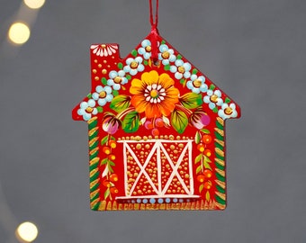 May include: Red wooden house ornament with a white and yellow floral design. The house has a white and gold window and a red roof.