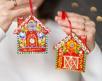 May include: Two red wooden ornaments shaped like houses with painted floral designs. The ornaments are decorated with colorful flowers and leaves. The text "The Andersons" is written on one of the ornaments.