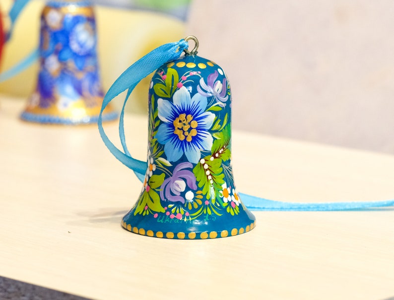 May include: A teal blue bell ornament with a floral design painted on it. The bell has a blue ribbon attached to the top.