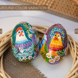 May include: Two hand-painted Easter eggs featuring a chicken design. One egg is green with a purple chicken, while the other is purple with a yellow chicken. Both eggs are decorated with white flowers and green leaves.