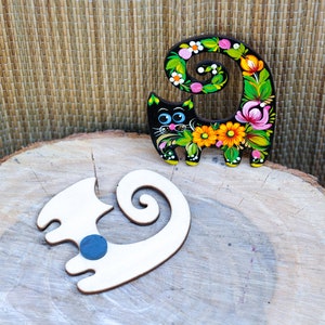 May include: Two wooden cat figurines, one painted black with colorful flowers and the other unpainted, on a wooden surface.