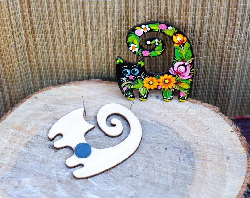 May include: Two wooden cat figurines, one painted black with colorful flowers and the other unpainted, on a wooden surface.