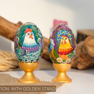 May include: Two hand-painted wooden Easter eggs with a rooster design, each on a gold stand. The eggs are decorated with colourful flowers and leaves. The rooster on the left egg is blue and purple, while the rooster on the right egg is yellow and red.