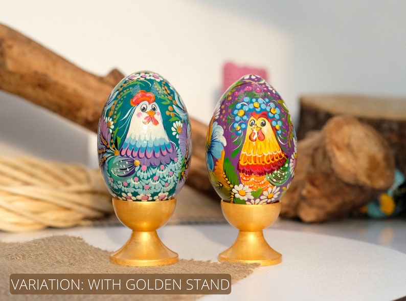 May include: Two hand-painted wooden Easter eggs with a rooster design, each on a gold stand. The eggs are decorated with colourful flowers and leaves. The rooster on the left egg is blue and purple, while the rooster on the right egg is yellow and red.