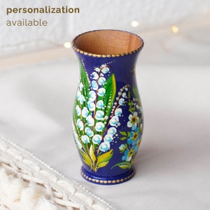 May include: A small, blue, hand-painted wooden vase with a floral design. The vase is decorated with white lily of the valley flowers and green leaves. The vase has a gold rim and a gold base.