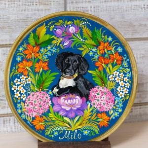 Puede incluir: Un plato de madera pintado a mano con fondo azul y un retrato de perro blanco y negro en el centro. El perro est&aacute; rodeado de flores y hojas coloridas. El plato est&aacute; firmado "Milo" en la parte inferior.
