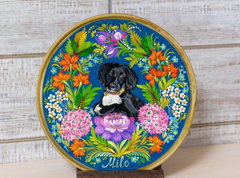 Puede incluir: Un plato de madera pintado a mano con fondo azul y un retrato de perro blanco y negro en el centro. El perro est&aacute; rodeado de flores y hojas coloridas. El plato est&aacute; firmado "Milo" en la parte inferior.