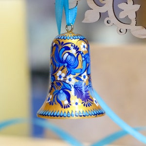Puede incluir: Una campana dorada con dise&ntilde;os florales azules y blancos pintados en ella. La campana est&aacute; colgada de una cinta azul.