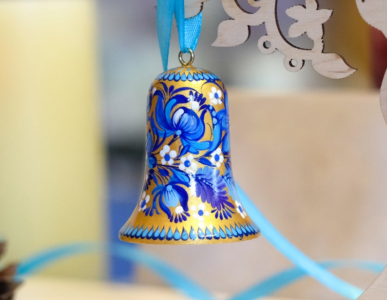 Puede incluir: Una campana dorada con dise&ntilde;os florales azules y blancos pintados en ella. La campana est&aacute; colgada de una cinta azul.