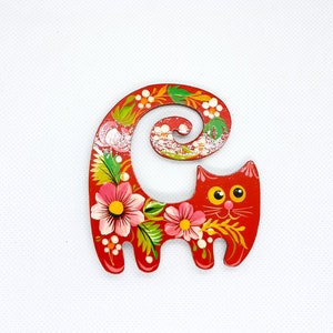 May include: A red wooden cat figurine with a curled tail and floral designs painted on it. The cat has yellow eyes and a white face.