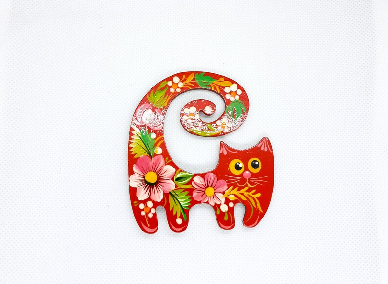 May include: A red wooden cat figurine with a curled tail and floral designs painted on it. The cat has yellow eyes and a white face.