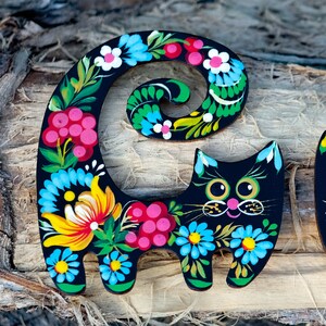 May include: Black wooden cat figurine with a curled tail painted with colorful flowers and swirls. The cat has a whimsical expression with large eyes and a pink nose.