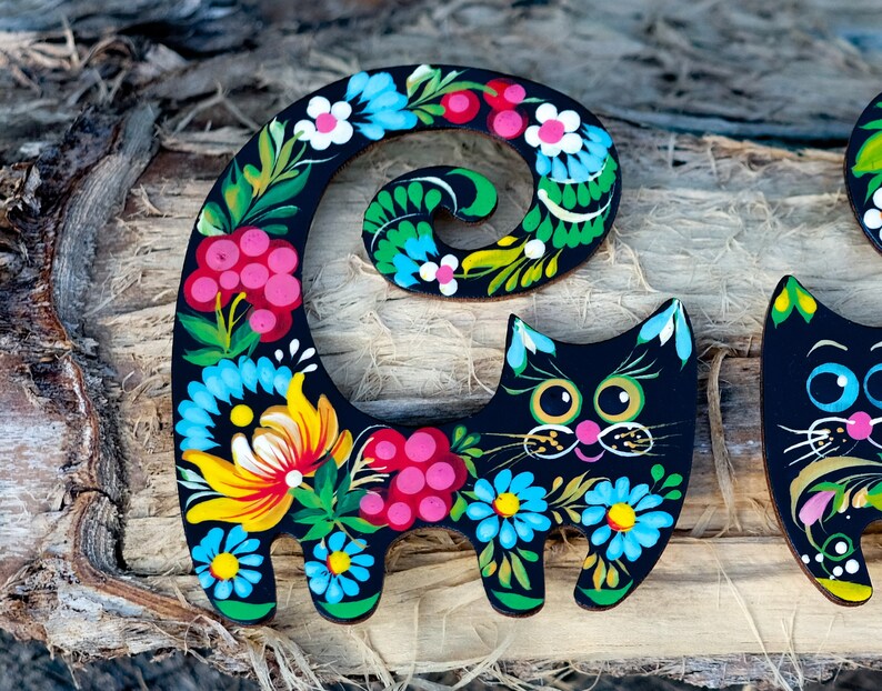 May include: Black wooden cat figurine with a curled tail painted with colorful flowers and swirls. The cat has a whimsical expression with large eyes and a pink nose.
