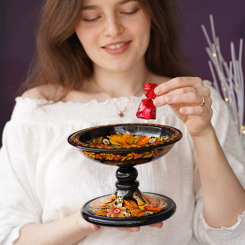 May include: A black and gold painted wooden bowl with a pedestal base. The bowl is decorated with floral and bird motifs. A person is holding a red wrapped candy over the bowl.